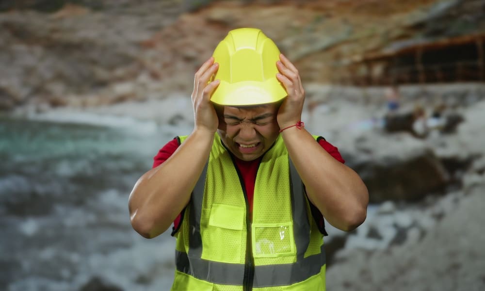 Man wearing safety gear looking stressed at seaside construction site with rocky background.